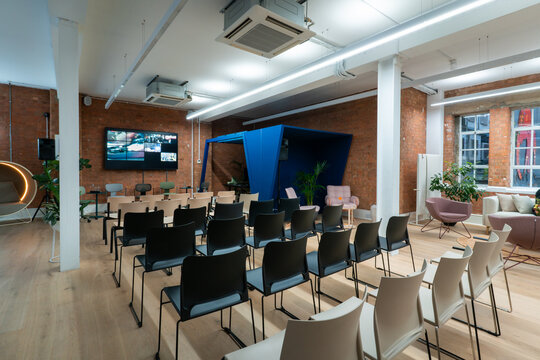View of a modern office meeting room with rows of black chairs, exposed brick walls, a large wall-mounted screen, and a blue acoustic pod in London, United Kingdom.