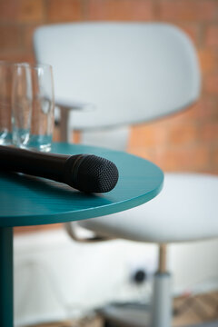 View of a black microphone resting on a teal round table next to two empty glasses, with a blurred white office chair and brick wall in the background in London, United Kingdom.