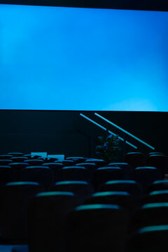 View of an empty cinema auditorium with rows of dark seats facing a large glowing blue screen and a small potted plant near the stage in London, United Kingdom.