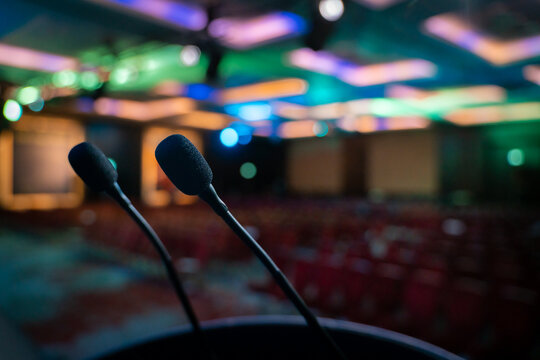 View of two microphones on a podium in a dimly lit conference room with colorful ambient lighting and rows of empty red seats in London, United Kingdom.