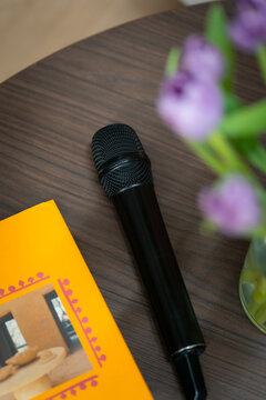 View of a black microphone resting on a dark wooden table next to a yellow book and a vase with purple flowers in London, United Kingdom.