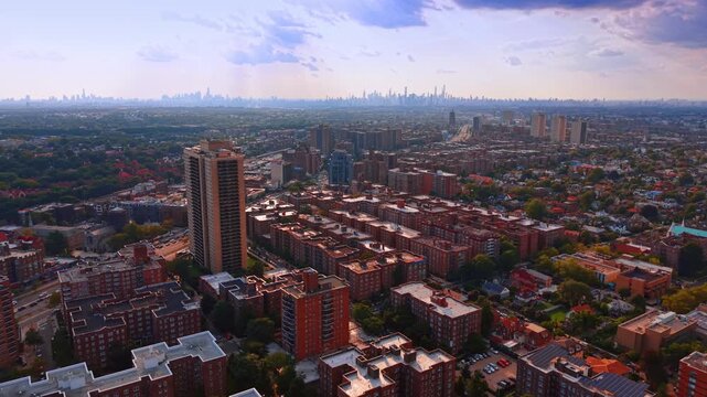 Apartment blocks stand beneath the distant Manhattan skyline. Warm light touches rows of brick buildings amid a green canopy under scattered clouds.