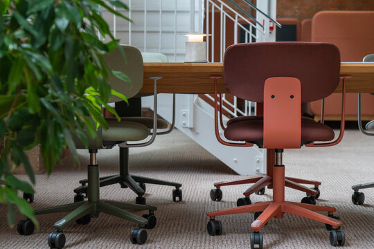 View of modern office chairs in green and terracotta colors arranged around a wooden table in a contemporary workspace with a green plant in the foreground, London, United Kingdom.