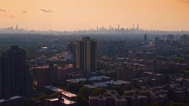 The Manhattan skyline appears beyond rows of apartment blocks. Dark rooftops and green trees fill the foreground below an orange sky with scattered clouds.