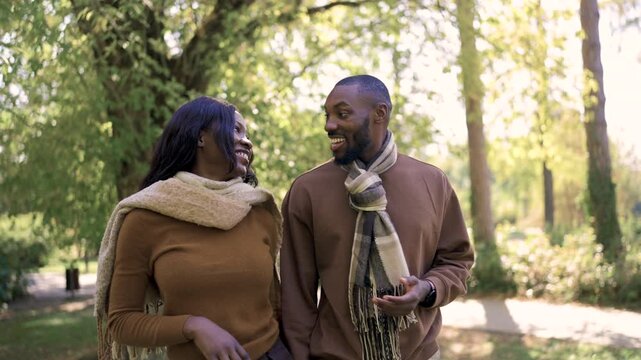 A joyful Black couple sits close together on a park bench, smiling warmly at each other during a peaceful afternoon outdoors. Their coordinated brown outfits and relaxed expressions convey love, compa