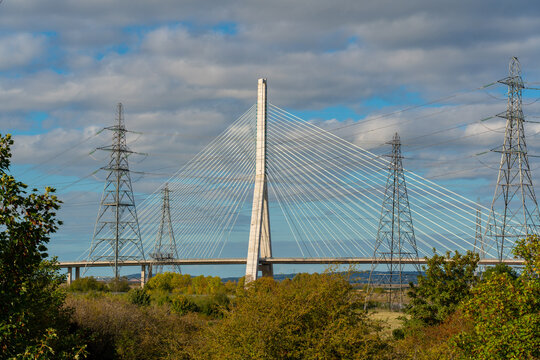 View of the cable-stayed Flintshire Bridge spanning the River Dee, framed by large electricity pylons and lush autumn foliage in Deeside, Wales, United Kingdom.