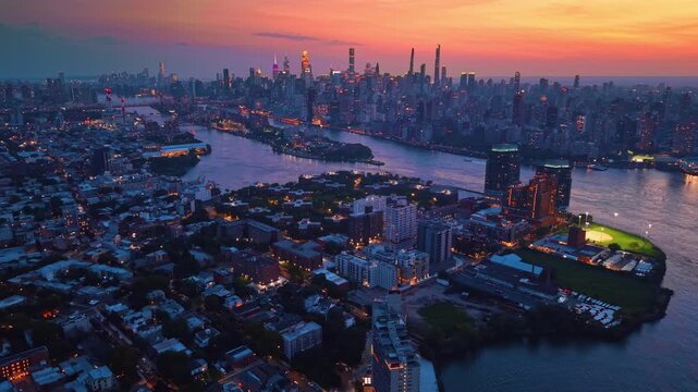 Manhattan towers rise beyond a New York waterfront neighborhood. Blue rooftops surround illuminated fields as orange light colors the river and clouded sky.