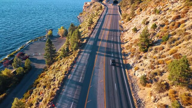 A car follows a lakeside highway beneath a rocky slope. Pine shadows stretch across gray lanes above blue Lake Tahoe water and a small parking area.