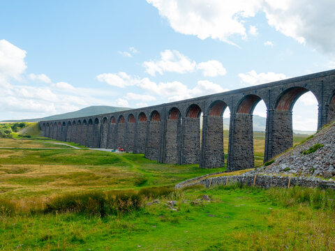 View of the historic Ribblehead Viaduct with its stone arches spanning the rolling green hills of the Yorkshire Dales under a bright sky in Carnforth, England, United Kingdom.