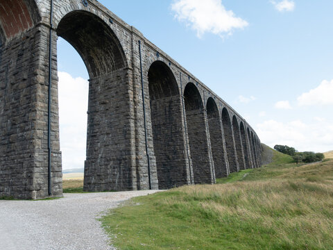 View of Ribblehead Viaduct, a massive stone railway bridge with multiple arches spanning a grassy landscape under a blue sky in Carnforth, England, United Kingdom.