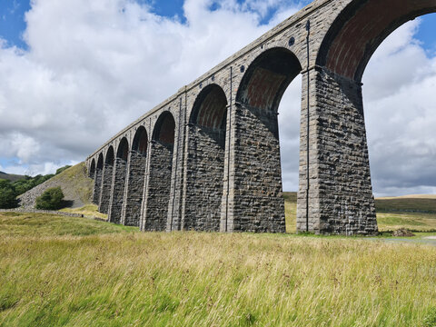 View of Ribblehead Viaduct, a historic stone railway bridge with multiple arches, set in a grassy landscape under a blue sky with clouds in Carnforth, England, United Kingdom.