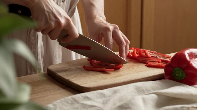 Woman's Hands Cutting Fresh Red Bell Pepper. Close-up of woman's hands slicing fresh red bell pepper into thin strips with a kitchen knife on a wooden cutting board
