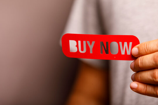 A person's hand holds a prominent red 'Buy Now' sign, urging immediate purchase.