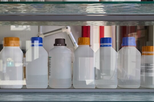 Laboratory plastic bottles with colorful screw caps standing in a row on a glass shelf. Group of chemical storage containers with transparent liquid or reagents in a science lab close up