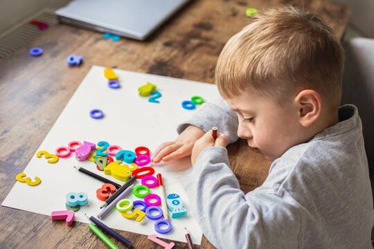 A cute little boy drawing with pencils and playing with colorful wooden numbers on paper during early childhood development at home.
