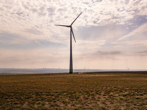 Aerial view of a large wind turbine standing in a vast dry field with a cloudy sky in the background near Tulcea, Tulcea, Romania.