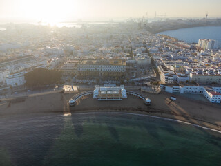 Aerial back-lit morning shot over La Caleta beach toward the ocean-encircled old town, with the sun rising dramatically behind the urban panorama. Cadiz, Andalusia, Spain. © nzatravel