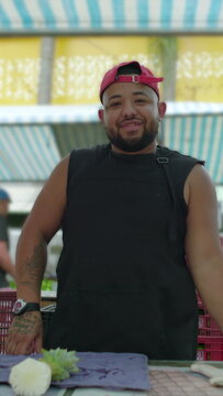 Smiling young Brazilian vendor in a backwards red cap and black vest at his street market stall