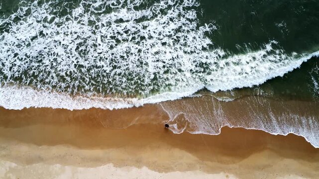 Aerial drone footage moving upward over a sandy beach as gentle ocean waves roll onto the shore, creating intricate white foam patterns and beautiful natural textures along the coastline.