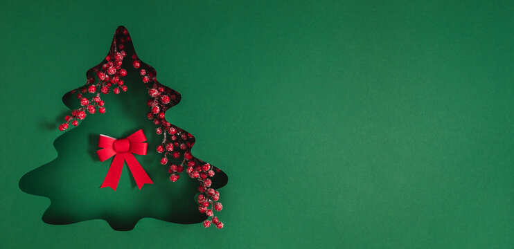 Minimalist Christmas background with a tree cutout in green paper, decorative red ribbon bow, and holly berries. Festive winter banner with copy space for New Year greeting card text.
