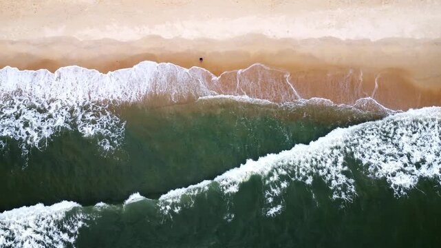 Aerial drone footage moving laterally to the right over a sandy beach as gentle ocean waves roll onto the shore, forming intricate white sea foam patterns against the deep green water. Top-down view s