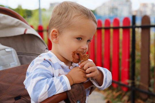 Side view of a little toddler eating a chocolate ice cream cone while sitting in a stroller outdoors.