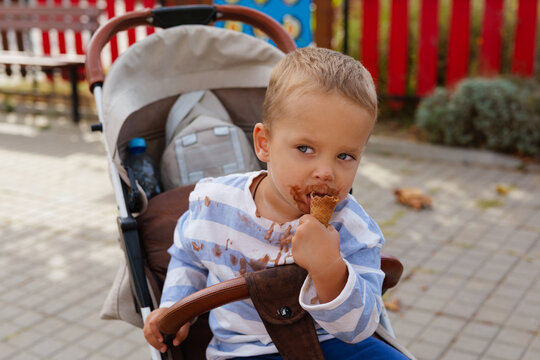 Cute messy toddler boy sitting in a stroller and eating a chocolate ice cream waffle cone outdoors.