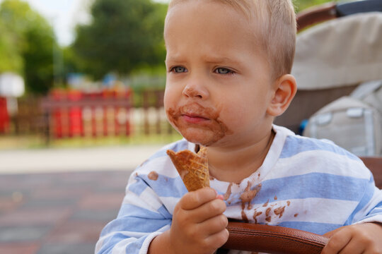 Close-up portrait of a messy toddler boy eating a chocolate ice cream cone in a stroller outdoors.