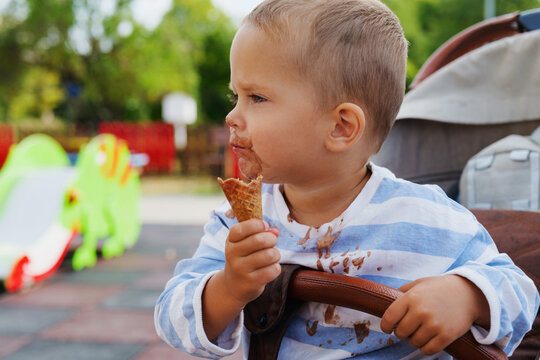 Little toddler sitting in a stroller outdoors and eating a chocolate ice cream cone with messy face and shirt.