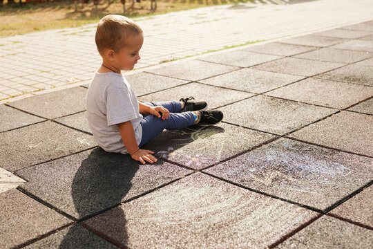 Little boy sitting on a playground rubber tile floor, looking at chalk drawings on the ground.