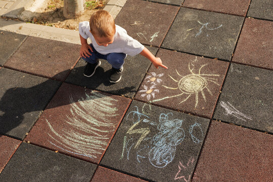 High angle view of a toddler boy pointing at a colorful chalk drawing of a sun on playground tiles.