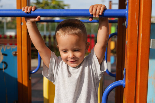 Cute little toddler boy holding onto a blue metal bar on a wooden playground structure outdoors.