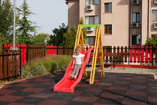 Little girl sliding down a red slide at an outdoor playground near a residential building.