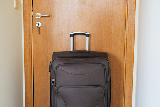 Front view of a brown fabric travel suitcase with telescopic handle standing in front of a wooden door with a key in the lock.