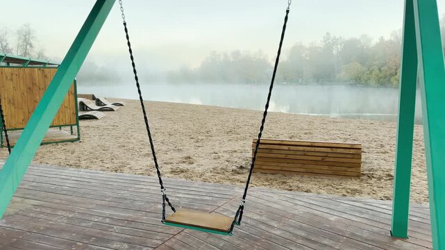 POV walkthrough of an empty lakeside beach park, shot on stabilizer. Smooth forward camera motion glides past a wooden swing facing a foggy lake in Pushkin, Russia.