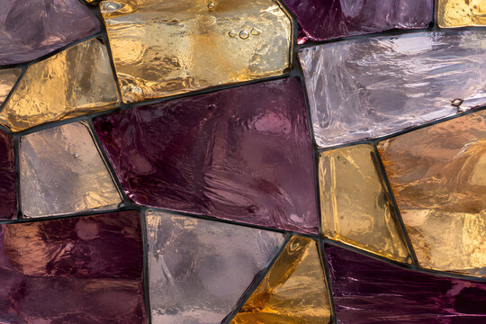 Close-up decorative mosaic of irregular translucent glass tiles in plum, amber and pale lilac tones with realistic surface imperfections.