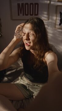 Vertical ugc portrait of young adult Caucasian woman pressing cold water bottle to forehead while talking to camera. Suitable for healthcare, summer heatwave, and wellness content