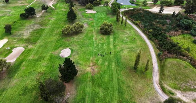 Aerial footage looks straight down over a golf course where a group of golfers stands on a striped fairway between sand bunkers and scattered trees. A curved service path and reed-lined water feature 