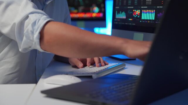 Financial data analyst typing on keyboard and looking at stock market charts and graphs on multiple computer screens at night