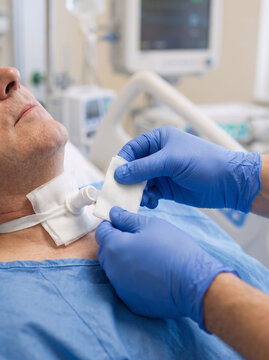 close-up of a trained healthcare professional carefully replacing a clean tracheostomy dressing on an adult patient, blue nitrile-gloved hands securing sterile gauze around the medical tube