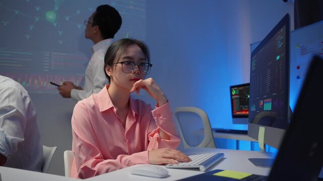 Young asian data analyst thinking and working on a computer during a team meeting in a modern office with blue neon light