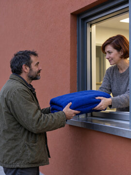 Kind woman giving blue blanket to homeless man through shelter window