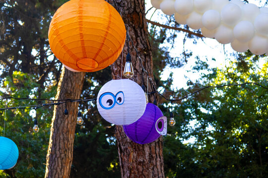 Colorful Halloween decorations featuring orange, white and purple paper lanterns with cartoon eyes, hanging light bulbs and white balloons among trees at an outdoor celebration.