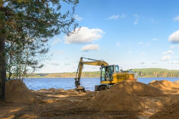 A yellow heavy excavator digger performing earthworks on a sandy beach by the river or lake. Construction, shore reinforcement, or beach improvement process on a sunny summer day. © Lexis_Jan