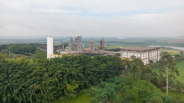 Aerial view of an industrial facility with a prominent white silo, metal structures, and large warehouses nestled among dense green trees near a river in Araras, Sao Paulo, Brasil.