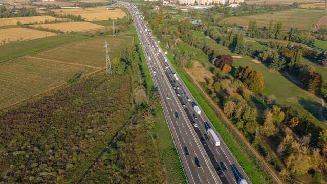 Aerial view of A50 Tangenziale Ovest di Milano with heavy traffic on the highway, surrounded by agricultural fields, green spaces, and trees in Milano, Lombardia, Italy.