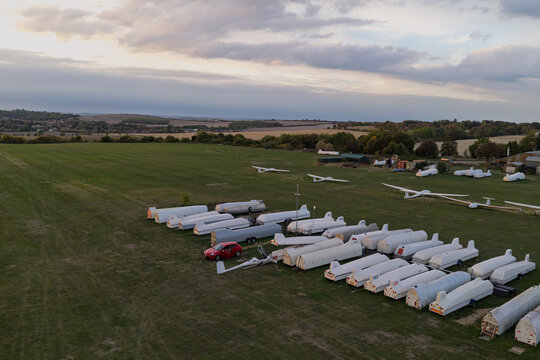 Aerial view of a glider airfield in the United Kingdom, featuring numerous gliders on grass, rows of white trailers, and a red car, set against a rural landscape under a cloudy sky.