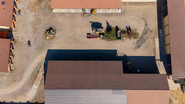 Aerial view of an agro farm featuring large buildings with solar panels, farm machinery, and vehicles parked on a dirt yard in Milan, Lombardy, Italy.
