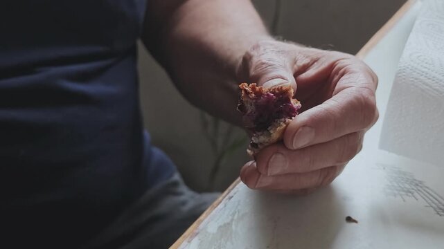 Close up of an elderly man's hands holding a piece of homemade blueberry muffin and drinking hot tea from a cup for breakfast