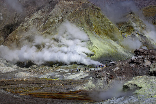 Aerial view of Whakaari White Island showing volcanic steam vents, sulfur deposits, and a group of tourists walking across the rocky terrain in Bay of Plenty Region, New Zealand.